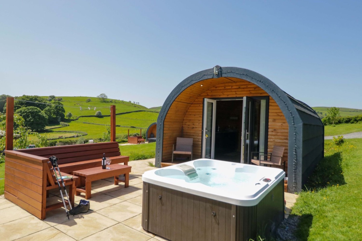 Glamping pod and hot tub on a timber deck with a bench, walking poles, and green Cumbrian hills