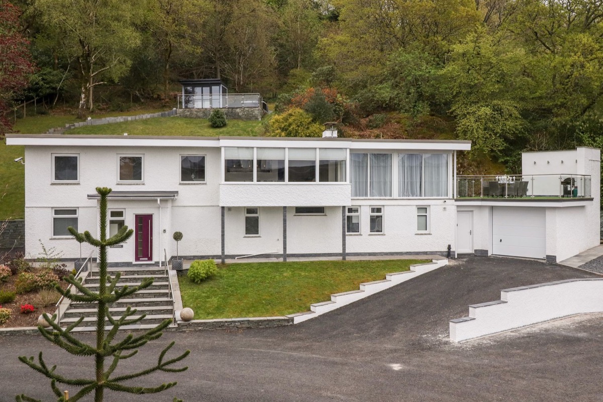 White modernist house set into a wooded hillside with terraced lawns and a driveway at Brantlea