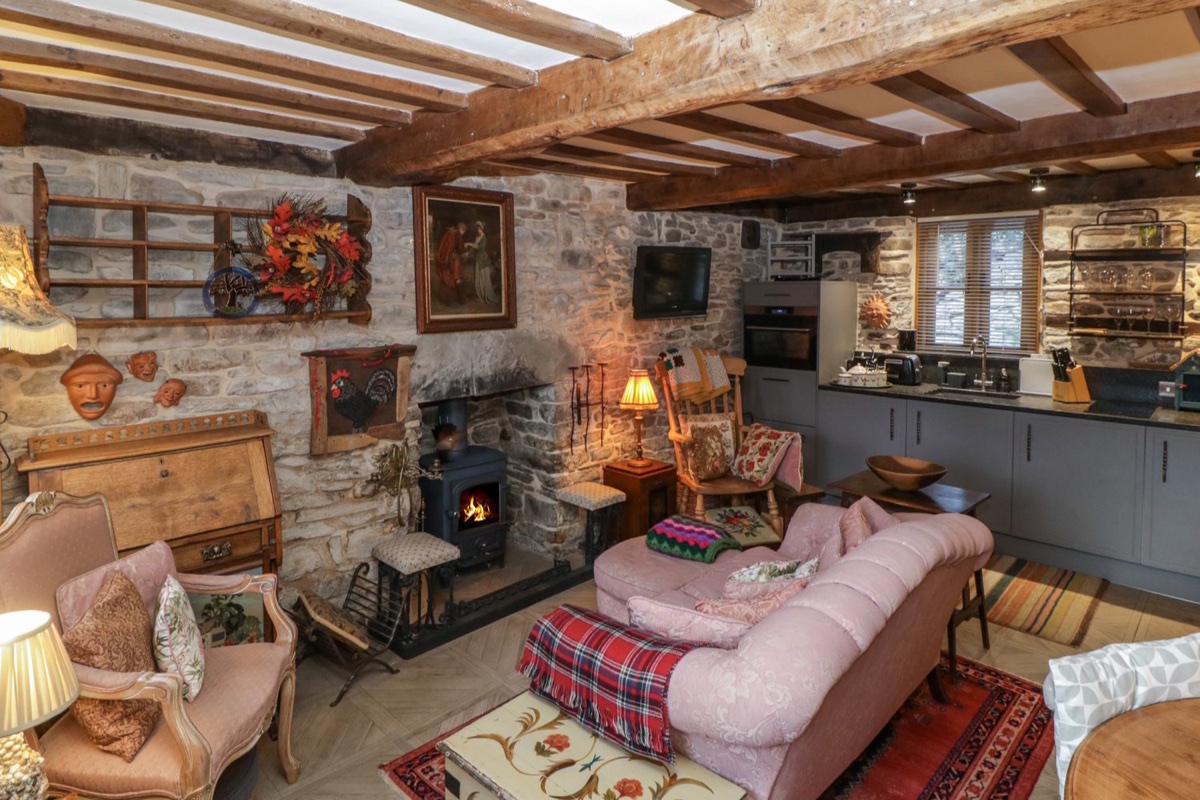 Living room with lit wood burner, stone walls, oak beams and pink sofa at Hoarders Cottage