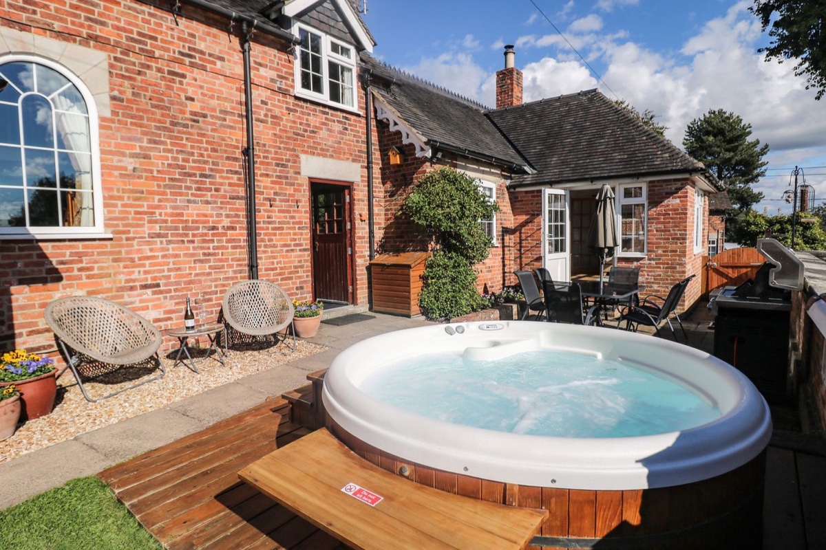 Hot tub on the deck in front of a red-brick cottage with patio seating at Orchard Cottage