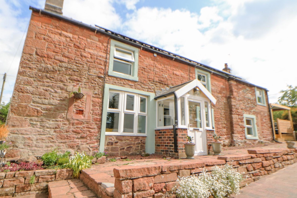 Red Penrith sandstone cottage with sage green window frames, white porch entrance, and garden flowers