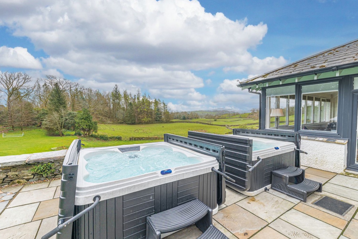 Two hot tubs side by side on a stone patio overlooking green fields and woodland near Windermere