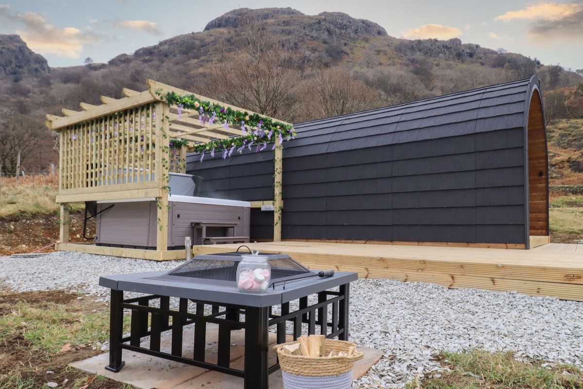 Black arched glamping pod with flower-draped pergola hot tub and fire pit against a rocky fell